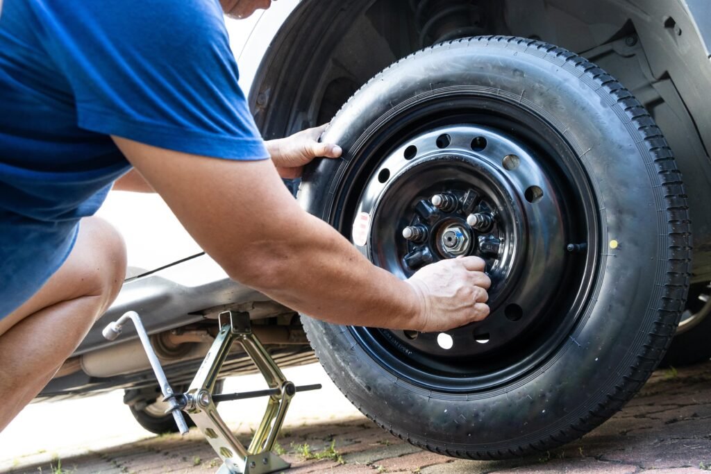 Person in a blue shirt mounting a spare tire onto a car's wheel hub while the vehicle is raised on a jack.