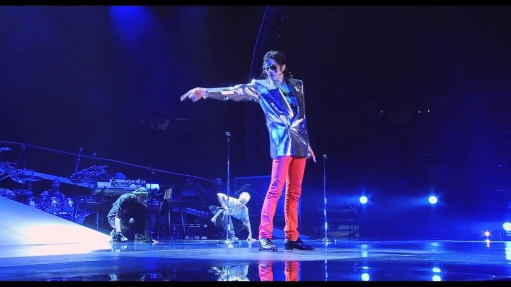 Michael Jackson performing on stage in a silver jacket and red pants, pointing toward the audience, with backup dancers and a band under blue stage lights.