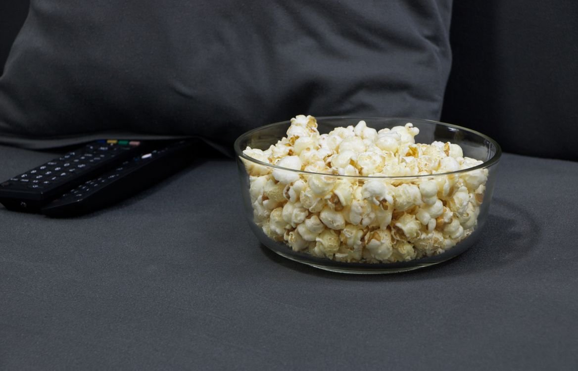 Glass bowl of popcorn next to TV remotes on a couch, ready for a Halloween movie night