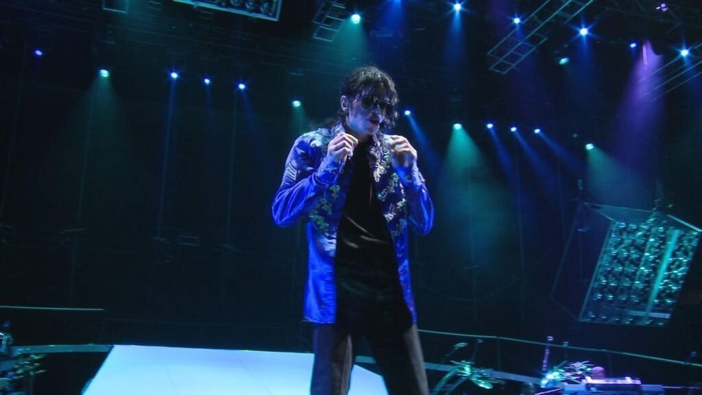 Michael Jackson performing on stage in a blue patterned jacket, holding a microphone, under blue and green stage lights.