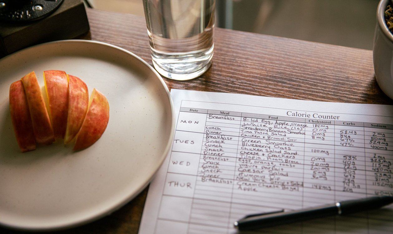 Handwritten weekly meal plan on paper next to a plate of sliced apple and a glass of water